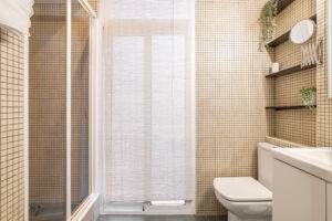 Nice bathroom with beige solid color tiles with a toilet, sink, walk-in shower, and spacious window.