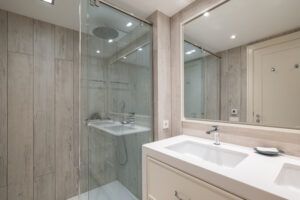 Spacious bathroom with bright artificial lighting, large mirror reflecting front door and pair of snow-white sinks built into light beige cabinet. Shower enclosure with blue glass splash guard.