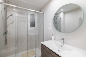 White ceramic tile bathroom with large round mirror on the wall reflecting the shower area with glass railing and metal fittings.